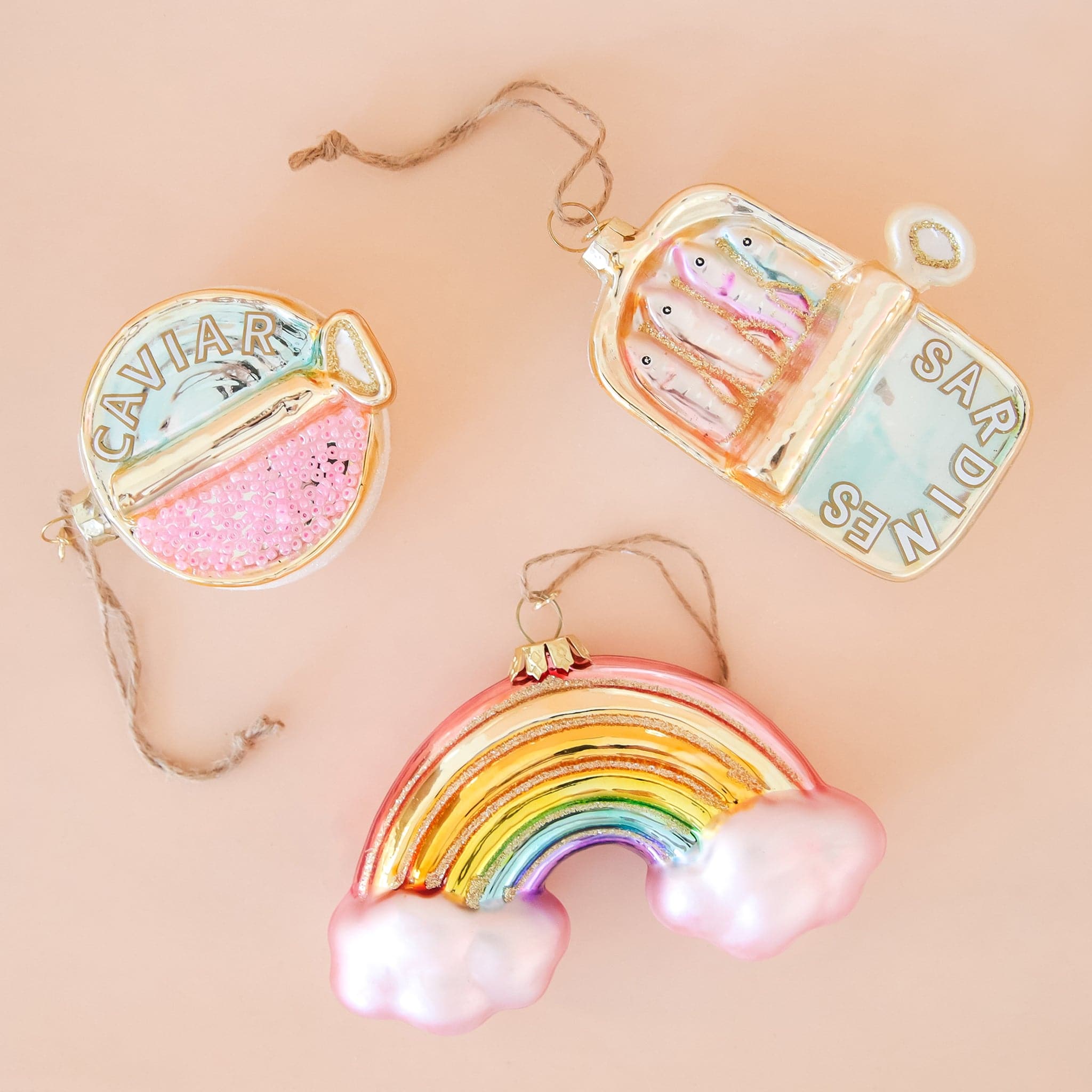 In front of a peachy background is three glass ornaments. On the bottom is a glass rainbow ornament. There is a light pink cloud on each end of the rainbow. Above it and on the right is a rectangular shaped ornament. One side is light blue with white text that reads ‘sardines.’ The other side has four colored fish. To the left is a circle ornament. The top half is light blue with white text that reads ‘caviar.’ The bottom half is covered with little pink beads. 