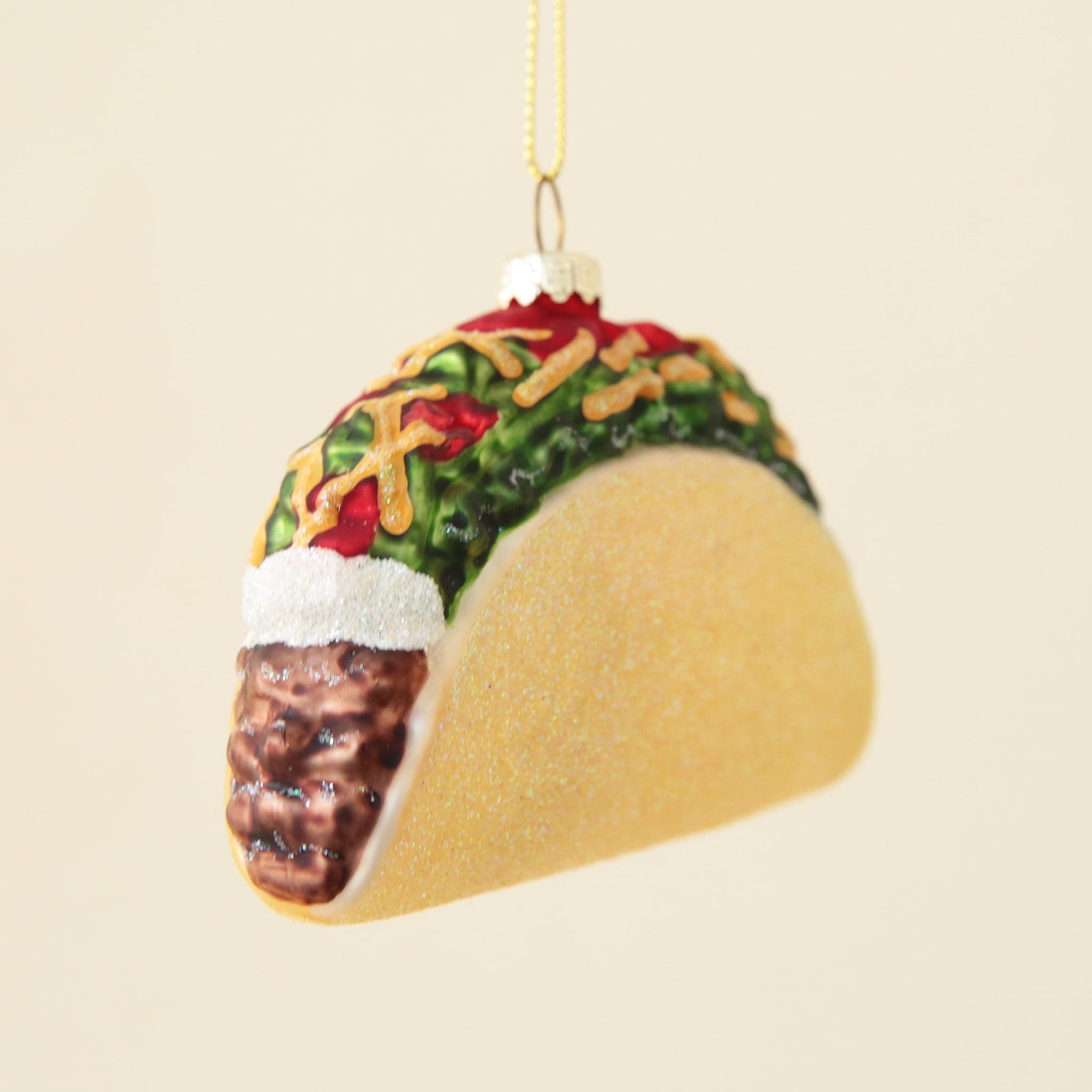 There’s a taco ornament hanging in front of a peachy background. The taco has a glittery yellow taco shell. Inside is brown glittery meat, white glittery sour cream, green glittery lettuce, red glittery tomatoes and yellow glittery cheese. The ornament is hanging by a gold hook and gold string.  