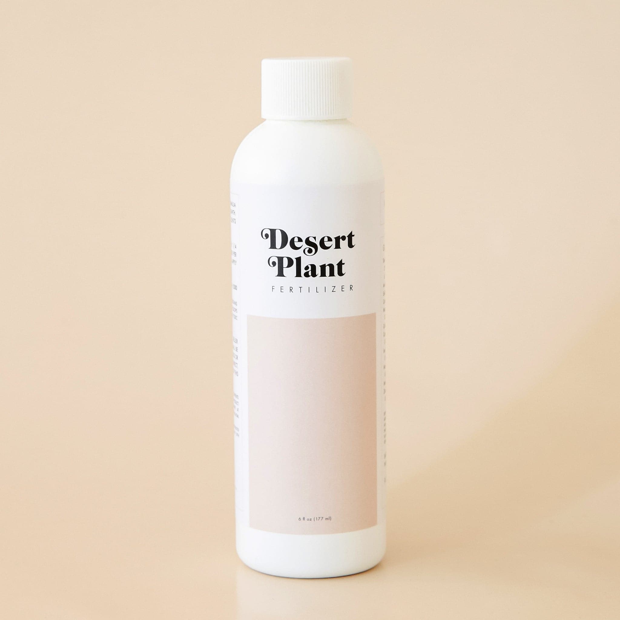 White twist top bottle labeled &#39;Desert Plant Fertilizer&#39; in black playful lettering. Below the label is a rectangular block of soft beige. The fertilizer sits in front of a solid sand colored background. 