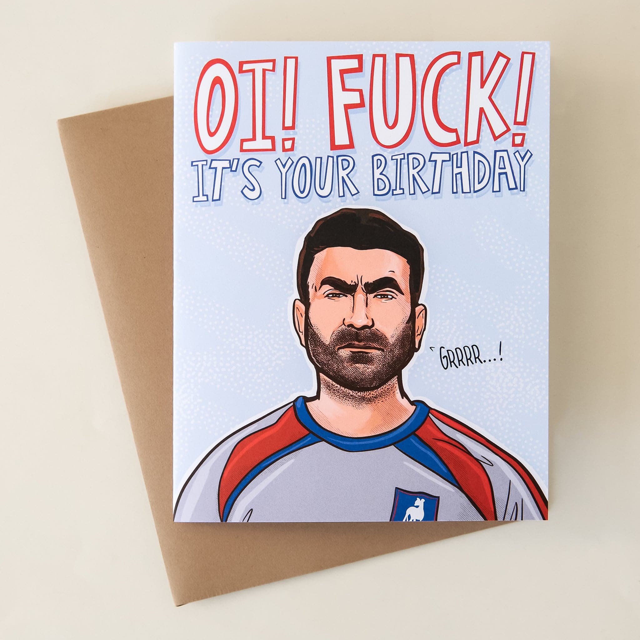 On top of a brown envelope is a light blue card. At top is white text that reads ‘OI! Fuck! It’s your birthday.’ Below is a drawing of the top half of a man wearing a grey, blue and red jersey. He has dark brown hair and brown beard. Next to him is black text that reads ‘grrrr…!'