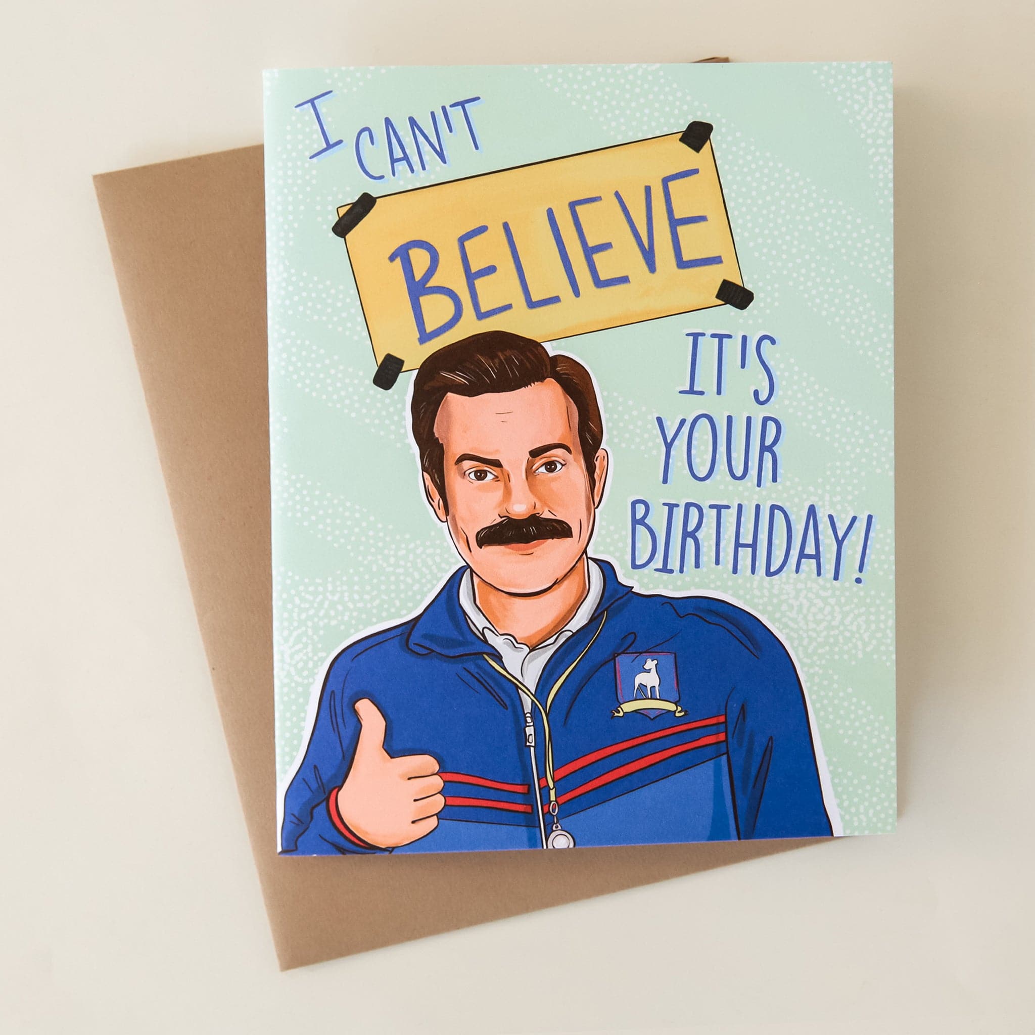 Against a white background is a mint colored card. At the bottom of the card is a drawing of Ted Lasso, a character from a tv show. The man has dark brown hair and a dark brown mustache. He has his right thumb up and is wearing a dark blue sports jacket. There is text above his head and next to his face. The blue text reads ‘I can’t believe it’s your birthday!'