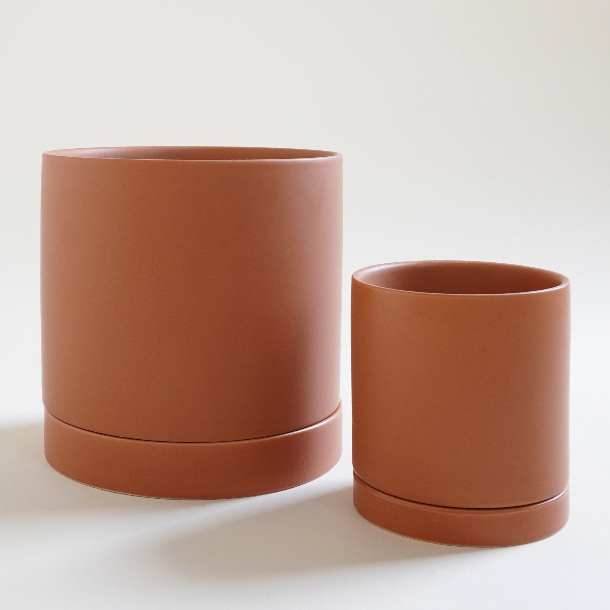 Two terracotta colored pots, one small and one large sit on a white ground