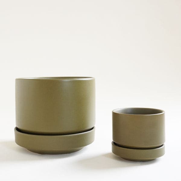In front of a white background is a round olive green pot with a matching tray that tapers at the bottom. To the right of the pot is another pot exactly like it just a lot smaller.