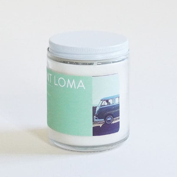 In front of a white background is a clear glass cylinder jar. The jar has a white top. On the front of the jar is a turquoise sticker. On the right side is a small picture of a VW bus in front of a blue sky. 