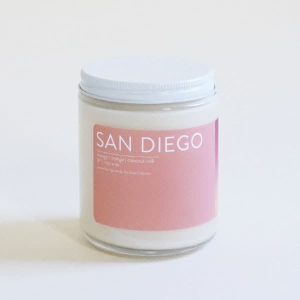 In front of a white background is a clear glass, round jar with a white metal lid. On the front is a pink sticker with white text at the top that reads ‘San Diego.&#39;