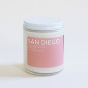 In front of a white background is a clear glass, round jar with a white metal lid. On the front is a pink sticker with white text at the top that reads ‘San Diego.'