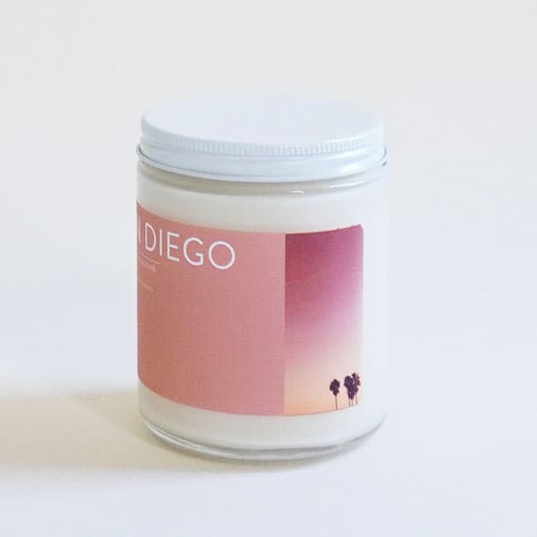 In front of a white background is a clear glass, round jar with a white metal lid. On the front is a pink sticker with white text at the top that reads ‘San Diego.’ On the left side of the sticker is a small picture of a dark pink and yellow sunset with a couple palm trees. 