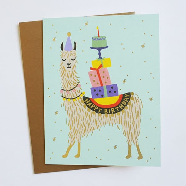 A light blue card with an illustration of a llama wearing a yellow happy birthday saddle and carrying a tower of multi colored wrapped gifts and a cake with a single candle on top. Also included is a coordinating envelope.