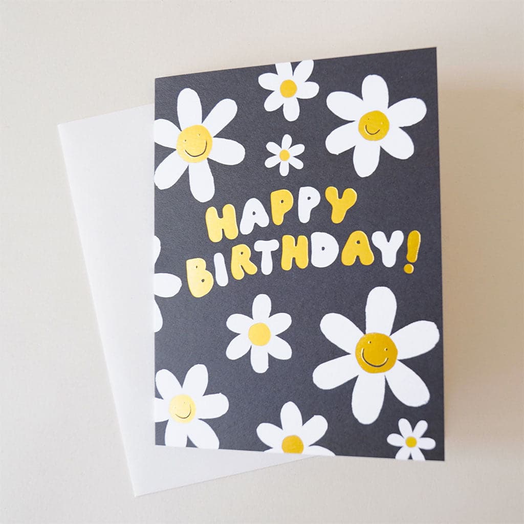 Black card reading 'Happy Birthday' in yellow and white bubble letters. The text is surrounded by white petaled, smiling daisies. The card is accompanied by a solid white envelope. 