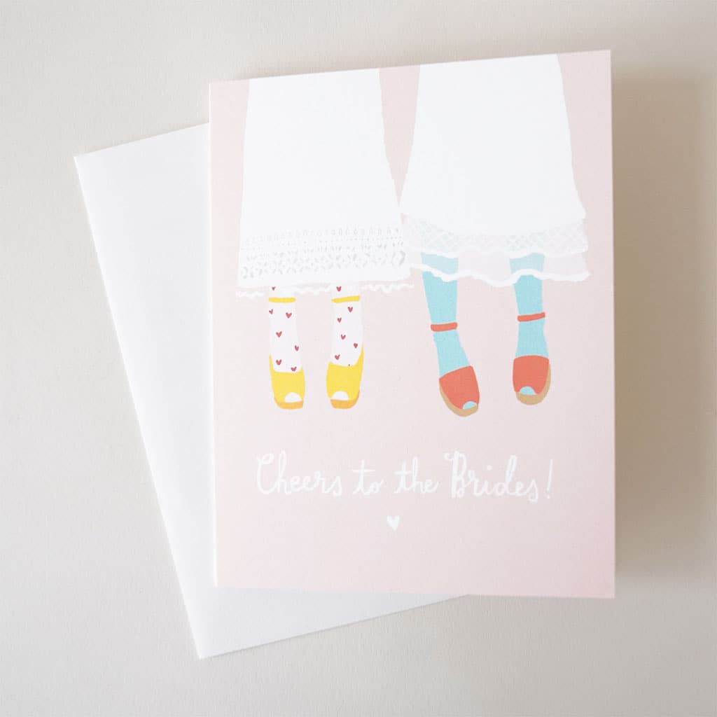 Soft pink card featuring two bottom halves of women. Both sets of legs are covered in a white midi skirt and colorful shoes and stockings. Below reads 'Cheers to the brides' in white cursive lettering. The card is accompanied by a solid white envelope. 