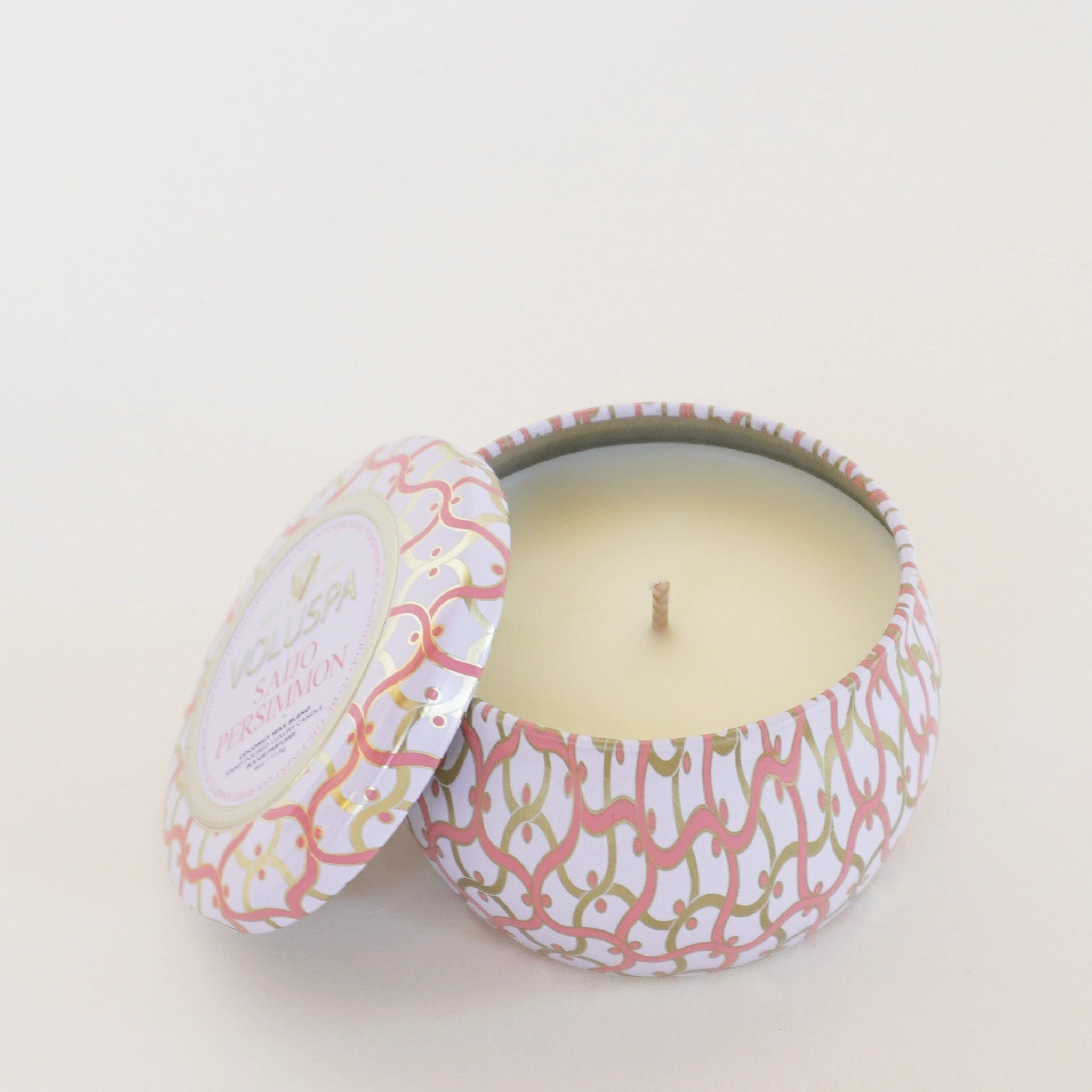 In front of a white background is a round tin candle. The tin is white with pink and gold swirly lines. Inside the tin is a white candle with a white wick in the center. There is a matching lid leaning against the left side of the candle.