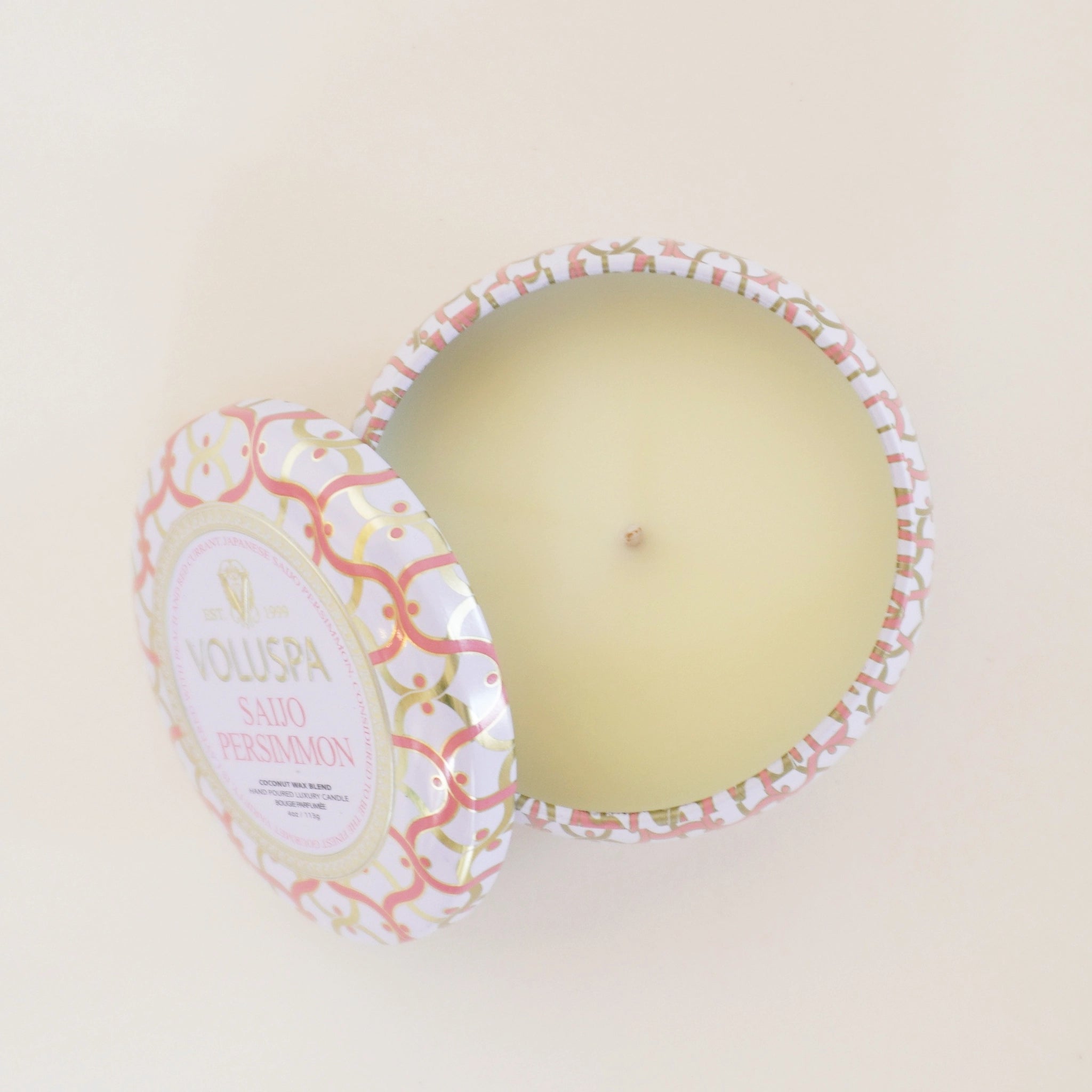 Birds eye view of a round tin candle. Inside is a white candle with a white wick in the center. The lid is leaning against the left side of the candle. The lid is whtie with pink and gold swirly lines. There is a white circle in the middle with gold text that reads ‘voluspa.’
