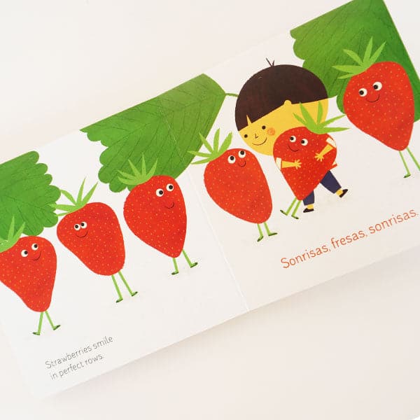 In front of a white background is an open book. Both pages are white. Across the two pages is a drawing of large green leaves at the top. There are red strawberries with green legs and smiley faces. They are walking across the pages. On the right page there is a child holding one of the strawberries. Under is orange text that reads ‘sonrisas, fresas, sonrisas.’ On the left page is black text that reads ‘strawberries smile in perfect rows.’