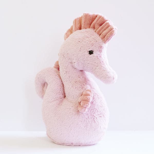 In front of a white background is the side view of a stuffed seahorse. The stuffed animal has a blush pink body and head with pink fins and mane. It has a black eye on the side of his head. 