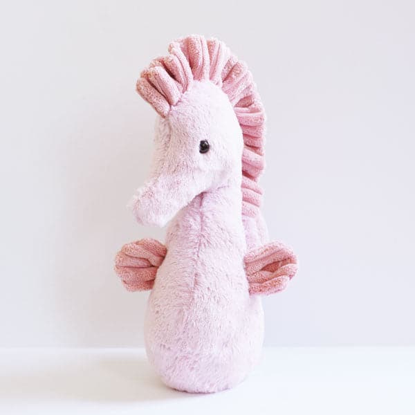 In front of a white background is the front view of a stuffed seahorse. His body is blush pink. He has a pink fin  on both sides of his body. There is a pink mane on the top of his head and down his back. On black eye is seen on the side of his head.