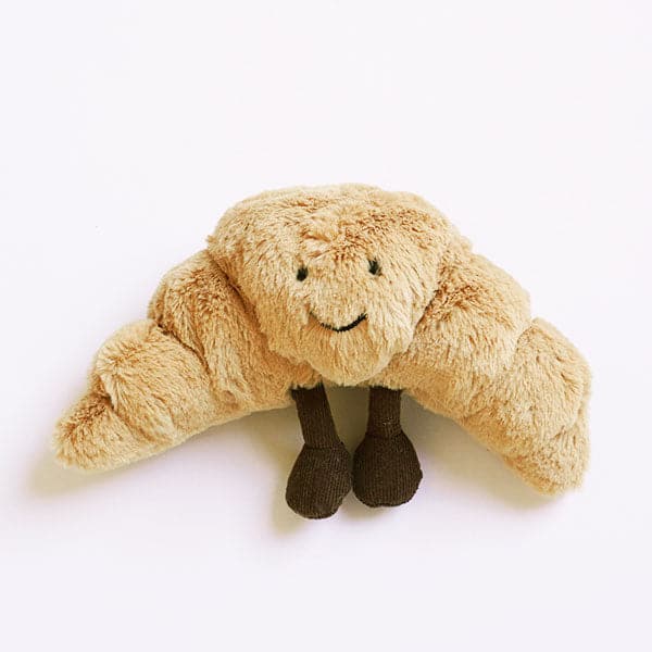 A soft stuffed animal in the shape of a croissant with smiley face and dark brown floppy legs.