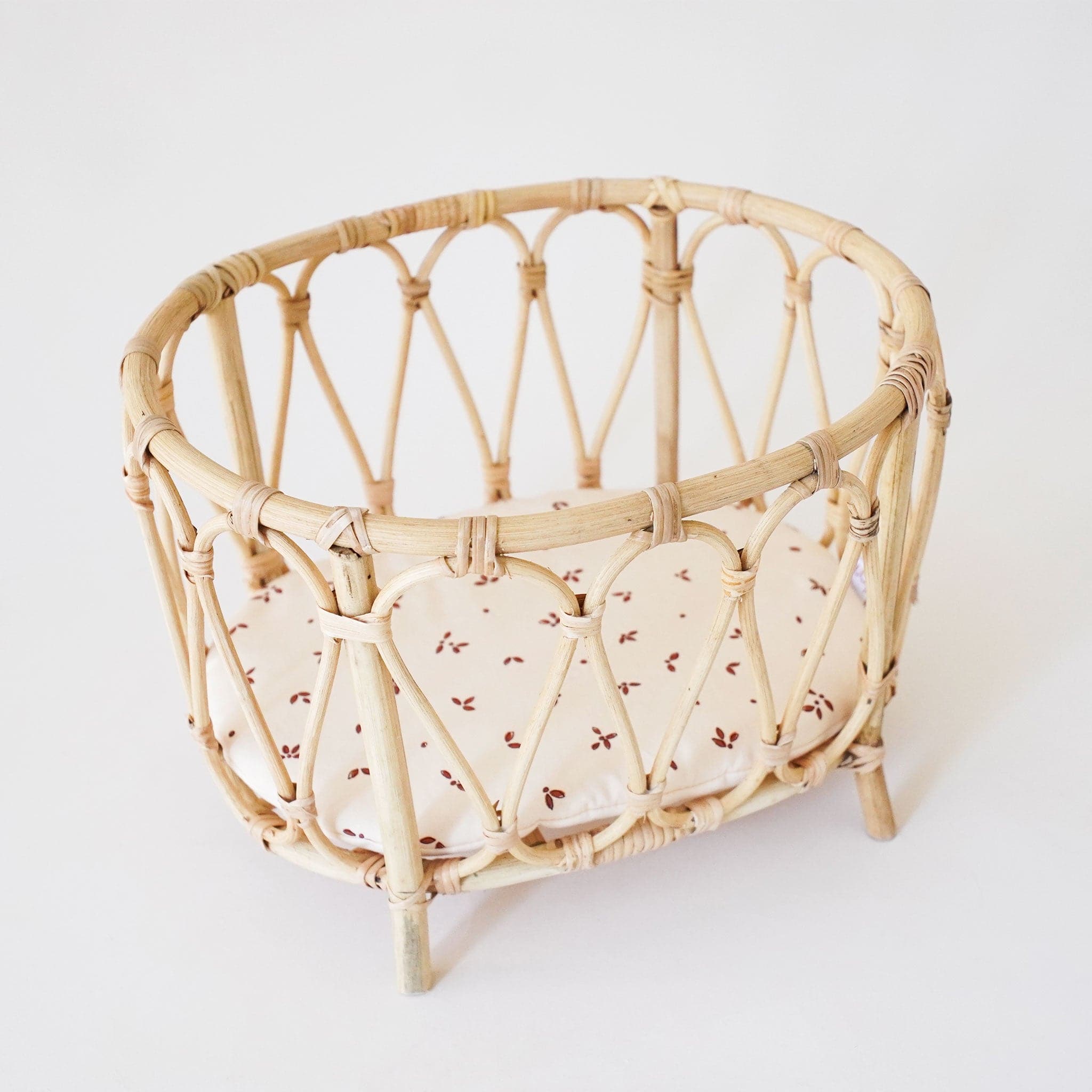 Doll playpen made of arched bamboo and rattan materials. The bed is elevated on four legs and the pad of the bed is ivory with small maroon detailing. Within the playpen sits a minikane doll wearing striped overalls. 