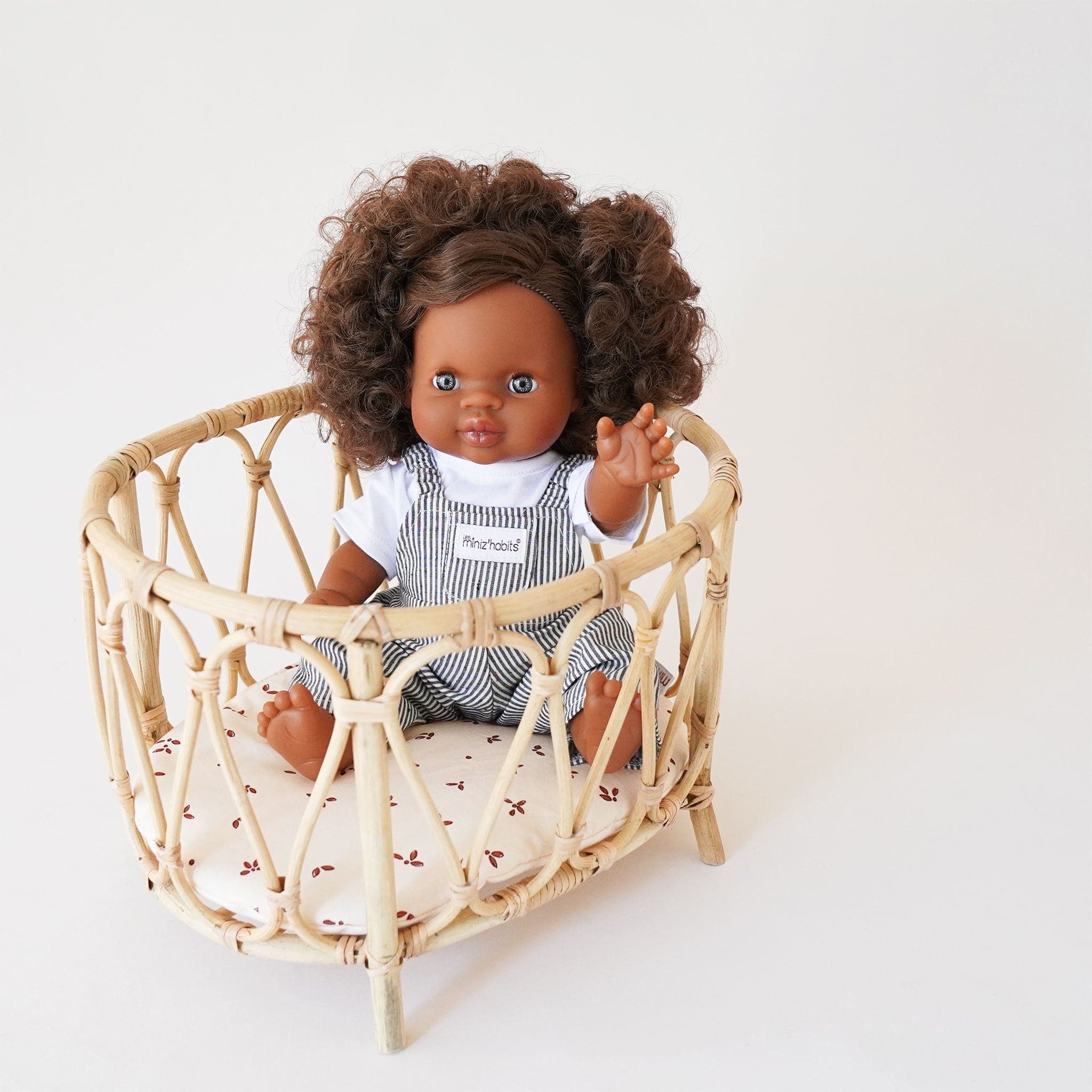 Doll playpen made of arched bamboo and rattan materials. The bed is elevated on four legs and the pad of the bed is ivory with small maroon detailing. Within the playpen sits a minikane doll wearing striped overalls. 