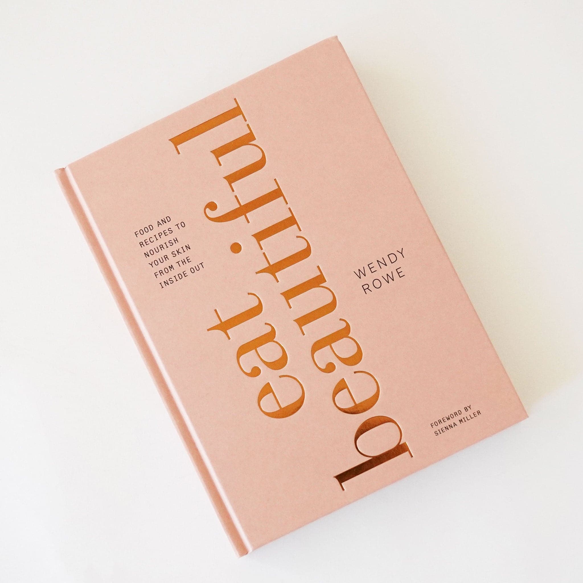 Cookbook with a soft pink hardcover that reads &#39;eat beautifully&#39; in large gold foil lettering facing perpendicular to the cover. In smaller lettering reads &#39;food and recipes to nourish your skin from the inside out&#39; in smaller lettering. 