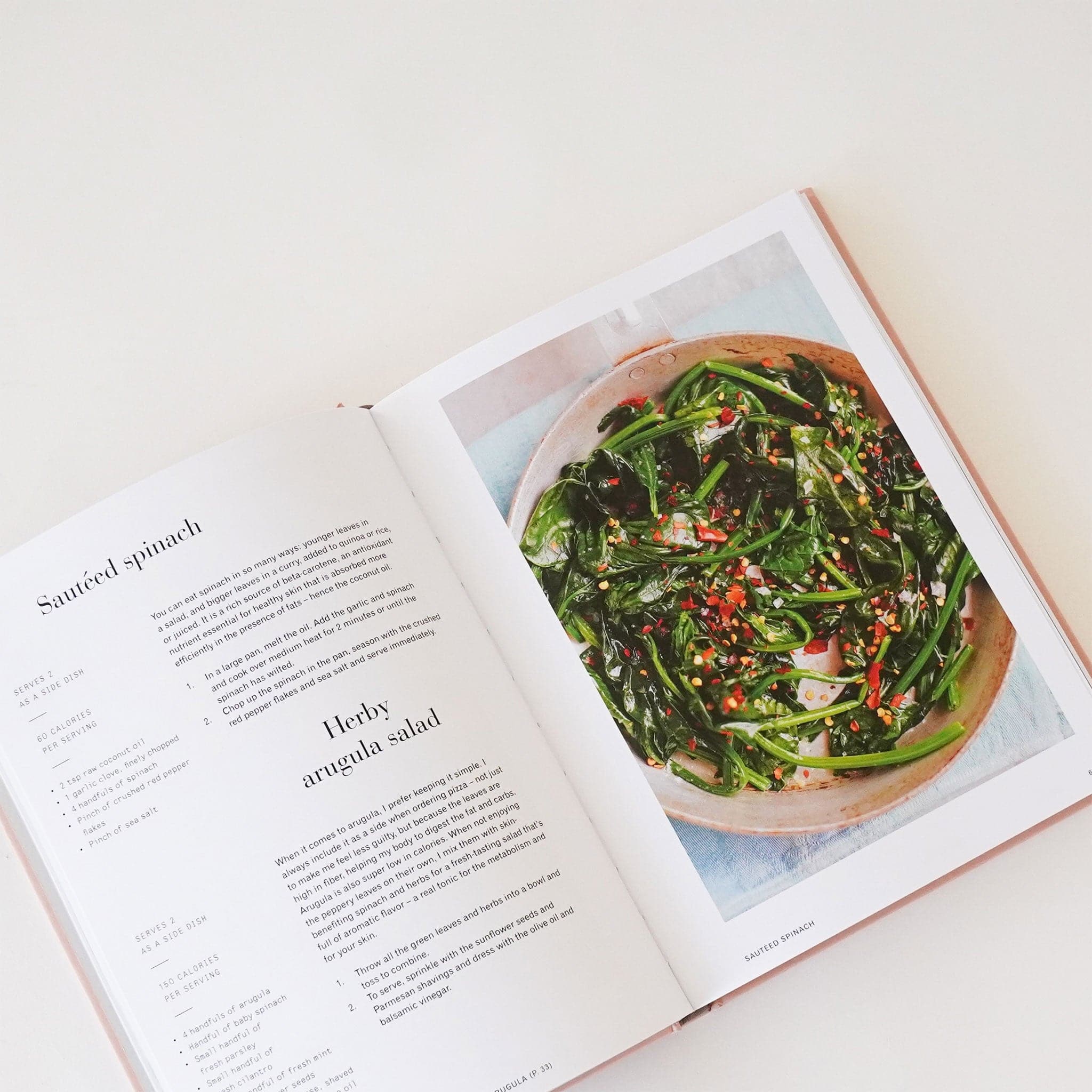 Cook book open to two pages. The left page lists a recipe for a sautéed spinach and Herby arugula salad. The right page features a plated vibrant green salad. 