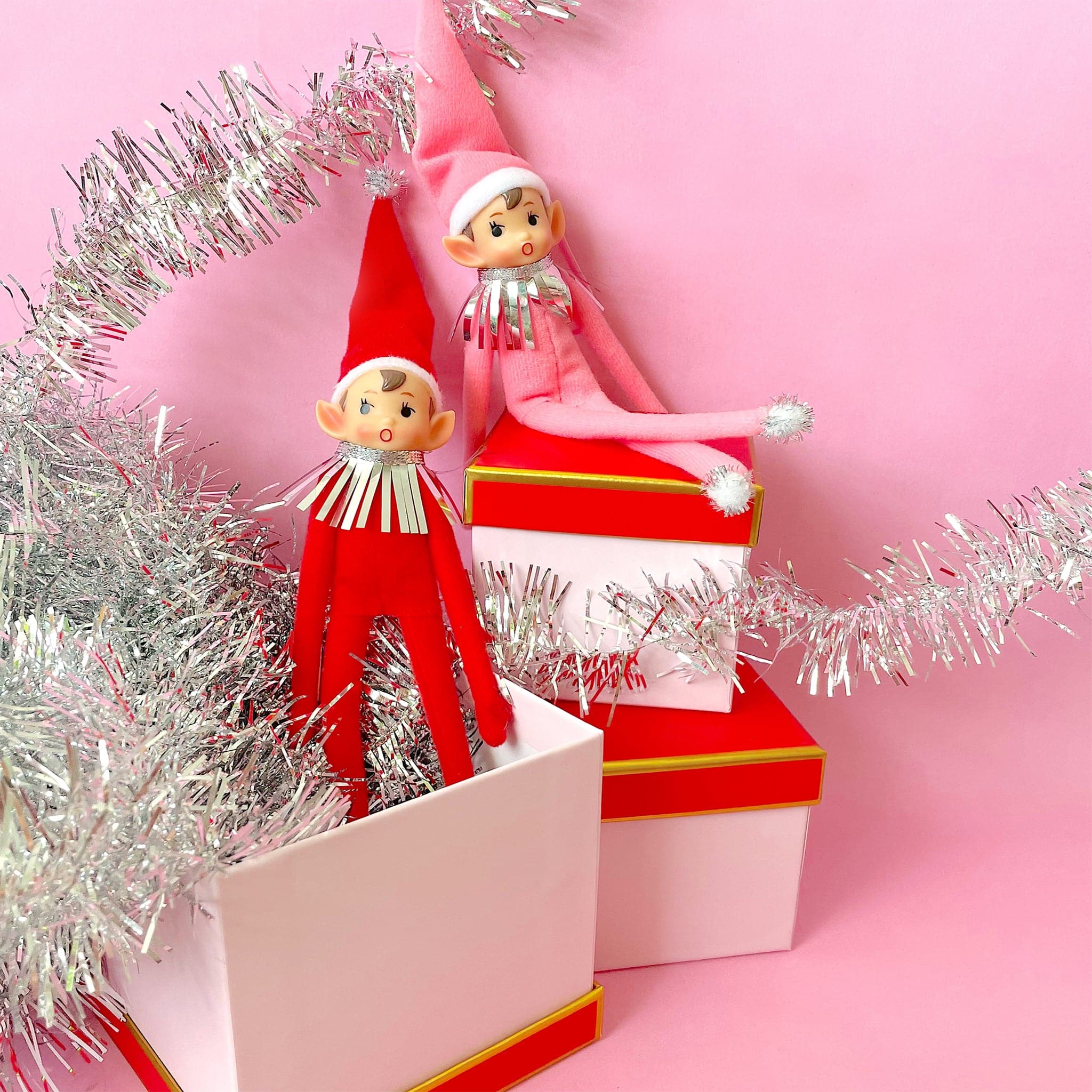 On a cream background is a red elf ornament with bendy arms and legs and a green loop for hanging and photographed next to the pink version.
