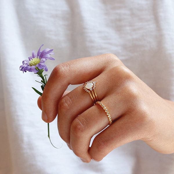 In front of a white background is a person’s hand holding a purple daisy. On the person’s middle finger is a stack of three rings. Each ring has a gold band. The top ring has an upward arch in the middle. The arch has three round diamonds on each side and a tall oval diamond in the middle. The middle band has a round white crystal in the middle with a small diamond on each side. The last band has a downward arch with seven small, round diamonds on the arch.