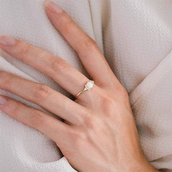 Worn on a models hand is a gold dainty ring with an opal stone with small CZ stones around it.