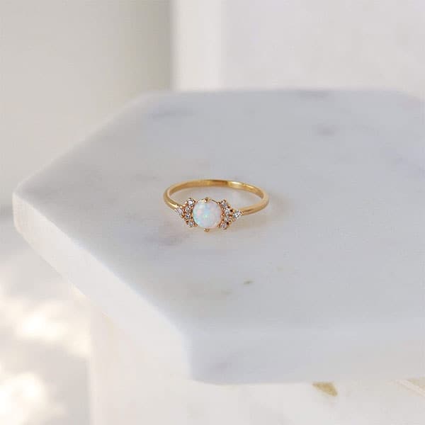 On a white background is a gold dainty ring with an opal stone with small CZ stones around it. 