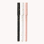 On a white background is a set of three pens, one black, one pink and one white that all have gold star and sun ray designs on them.