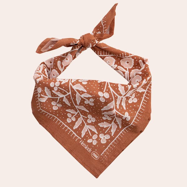 Against a white background is a dark terracotta bandana. The bandana is split into four triangles with white and pink flowers and leaves. The bandana is folded and tied at the top. 