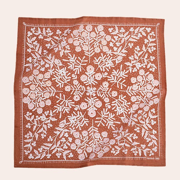 Against a white background is a dark terracotta bandana. The bandana is split into four triangles with white and pink flowers and leaves. 