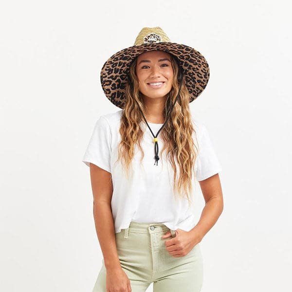 In front of a white background is a woman wearing a white t-shirt and tan pants. On her head she is wearing a wide brim, straw hat. The brim of the hat has a black and brown cheetah print border. It also has a black drawstring.