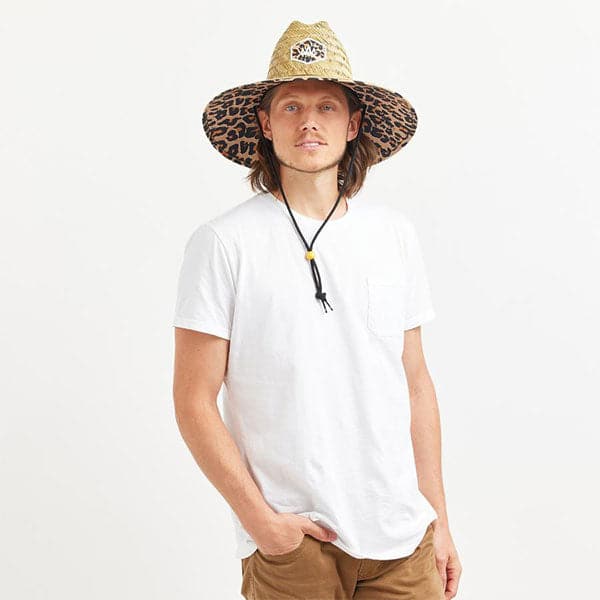 In front of a white background is a man wearing a white t-shirt and tan pants. On his head he is wearing a wide brim, straw hat. The brim of the hat has a black and brown cheetah print border. It also has a black drawstring.