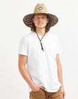 In front of a white background is a man wearing a white t-shirt and tan pants. On his head he is wearing a wide brim, straw hat. The brim of the hat has a black and brown cheetah print border. It also has a black drawstring.