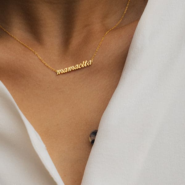 Zoomed in photo of a woman&#39;s neck. She is wearing a necklace that reads &quot;mamacita&quot; in lowercase script. A thin chain is connected on each side of the word.