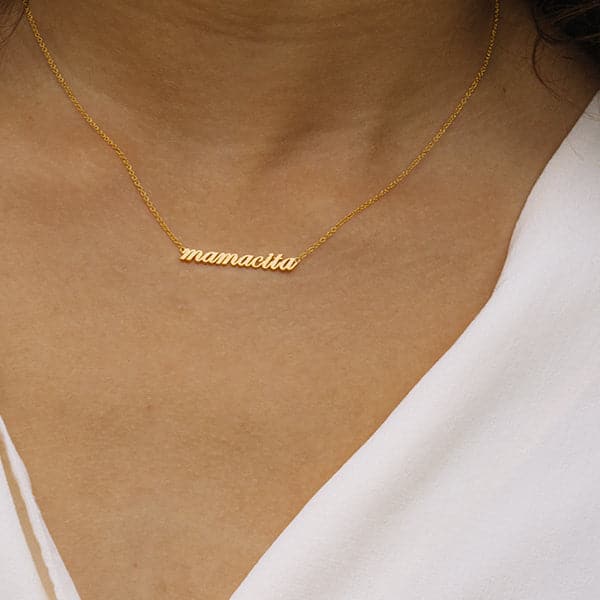 Zoomed in photo of a woman&#39;s neck. She is wearing a necklace that reads &quot;mamacita&quot; in lowercase script. A thin chain is connected on each side of the word.