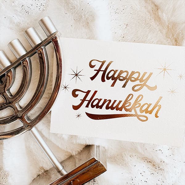 ivory greeting card that reads happy Hanukkah in cursive gold foil and surrounded by six starbursts on a soft furry background with a silver menorah styled next to it.
