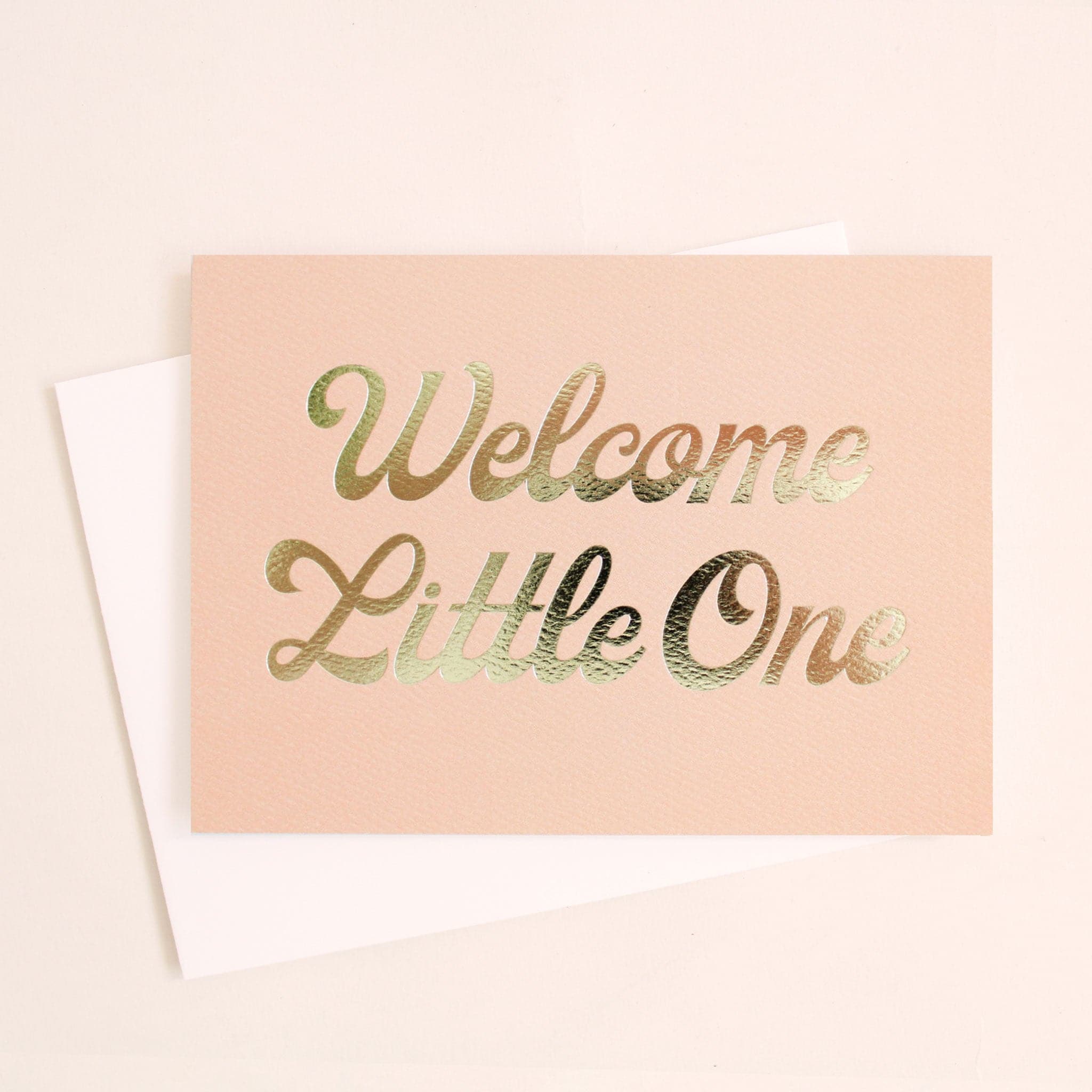 This card is a cloud peach color and reads 'Welcome Little One' in gold foil cursive lettering. The text takes up majority of the card. The card is accompanied by a solid white envelope.