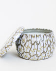 Against a white background is the side view of a round tin candle with a matching lid leaning against the left side of the candle. The candle and the lid are white with a gold and silver design. Inside the tin is a white candle. 