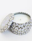 Against a white background is the side view of a round tin candle with a matching lid leaning against the left side of the candle. The candle and the lid are white with a gold and silver design. Inside the tin is a white candle. 