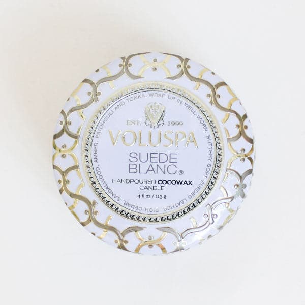 Against a white background is the birds eye view of a round, tin candle. The lid is white with a gold and silver design. In the center is a white circle with a gold and silver border. In gold text it reads ‘Voluspa.’ Below is black text that reads ‘suede blanc.’ 