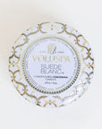 Against a white background is the birds eye view of a round, tin candle. The lid is white with a gold and silver design. In the center is a white circle with a gold and silver border. In gold text it reads ‘Voluspa.’ Below is black text that reads ‘suede blanc.’ 