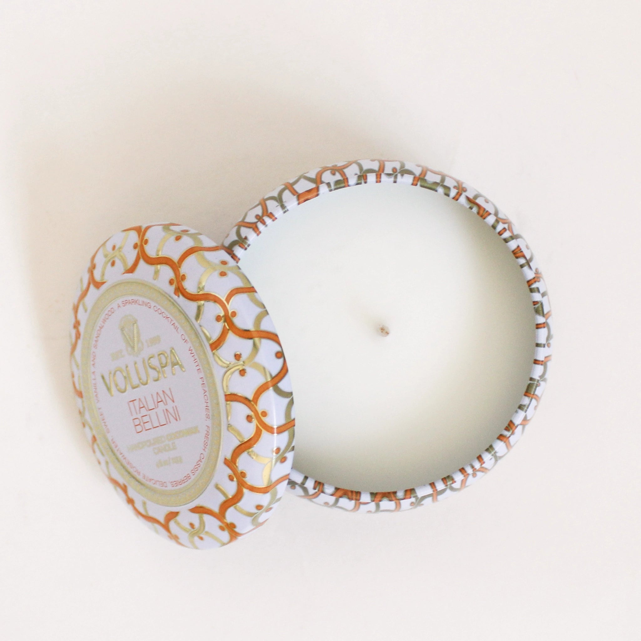 On a cream background is a orange and white tin that contains a single wick candle along with a circle label in the center of the lid that reads, "Voluspa Italian Bellini".