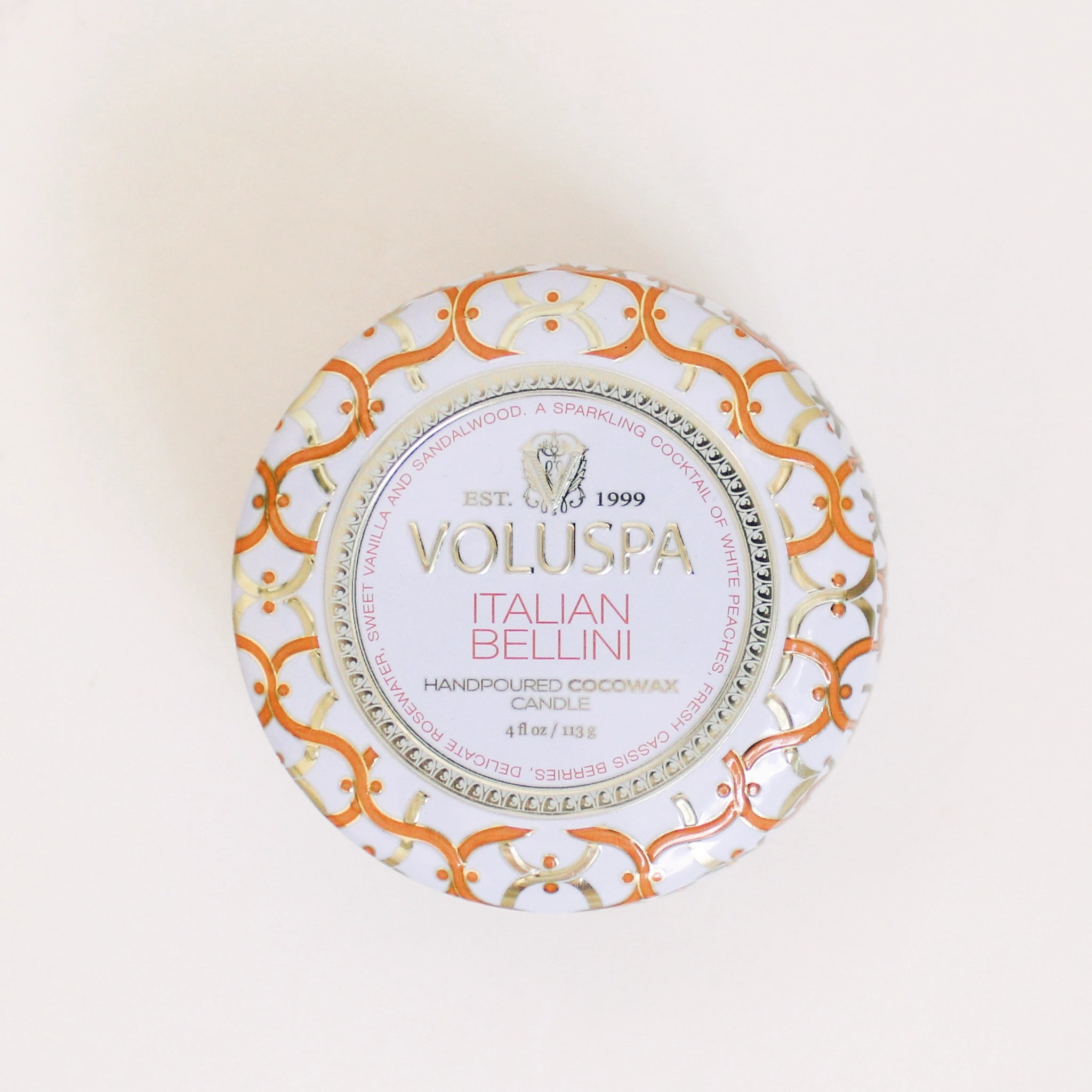 On a cream background is a orange and white tin that contains a candle along with a circle label in the center of the lid that reads, "Voluspa Italian Bellini".