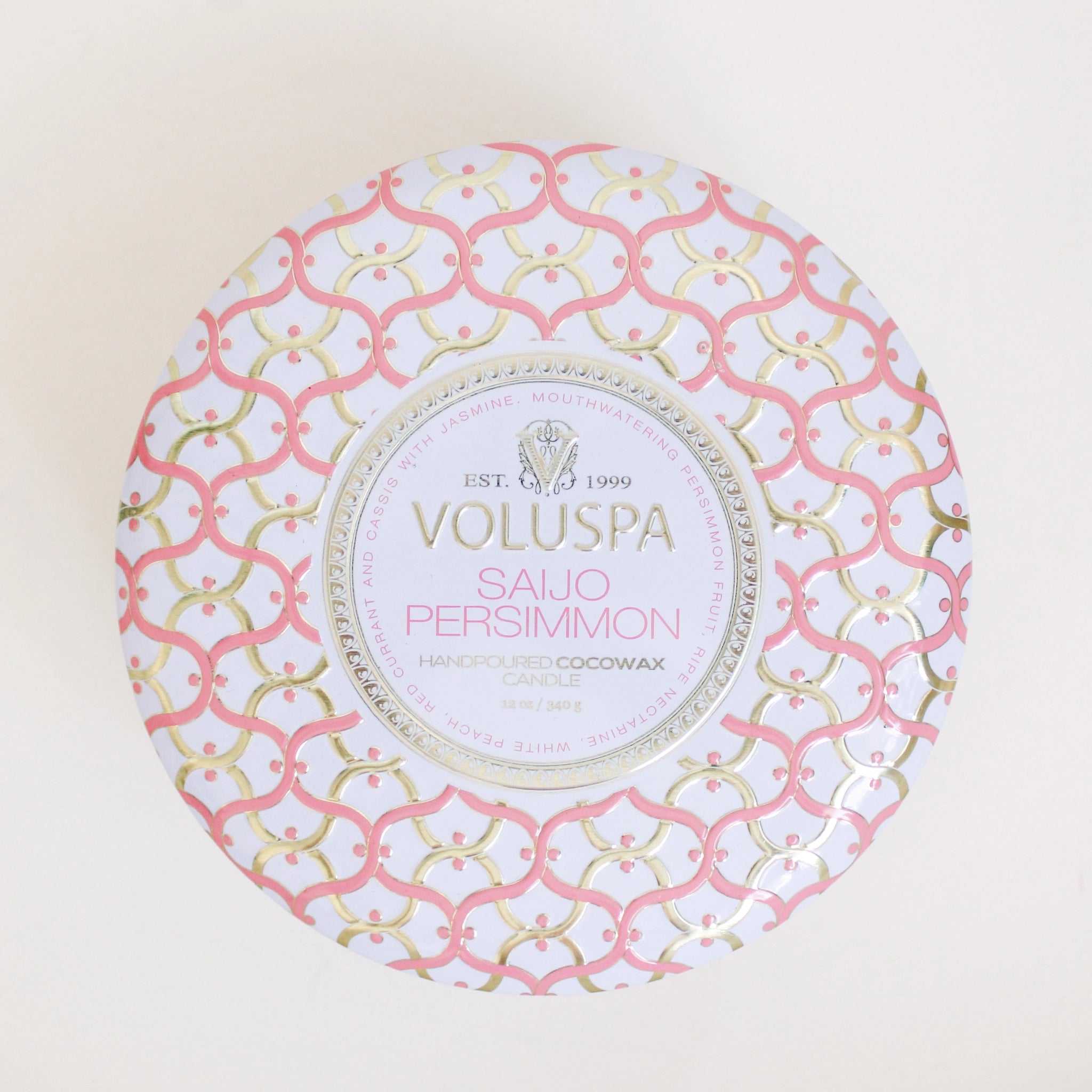 Birds eye view of the top of a round tin candle. The lid is white with pink and gold swirly lines on it. In the middle is a white circle with gold text that reads ‘voluspa.’ Below is pink text that reads ‘saijo persimmon.&#39;