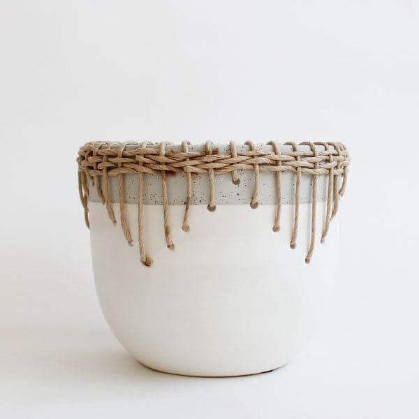 In front of a white background is a matte white, cylinder pot. There is a light gray border at the top of the pot. There is a light brown, wicker border over the gray border. The wicker border comes down the sides of the pot. 