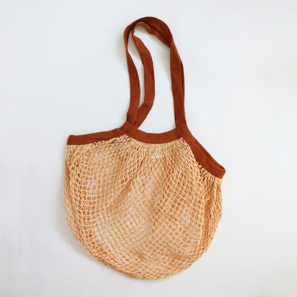 On a cream background is an orange and brown woven mesh shopping bag with a diamond pattern and two over the shoulder handle straps.
