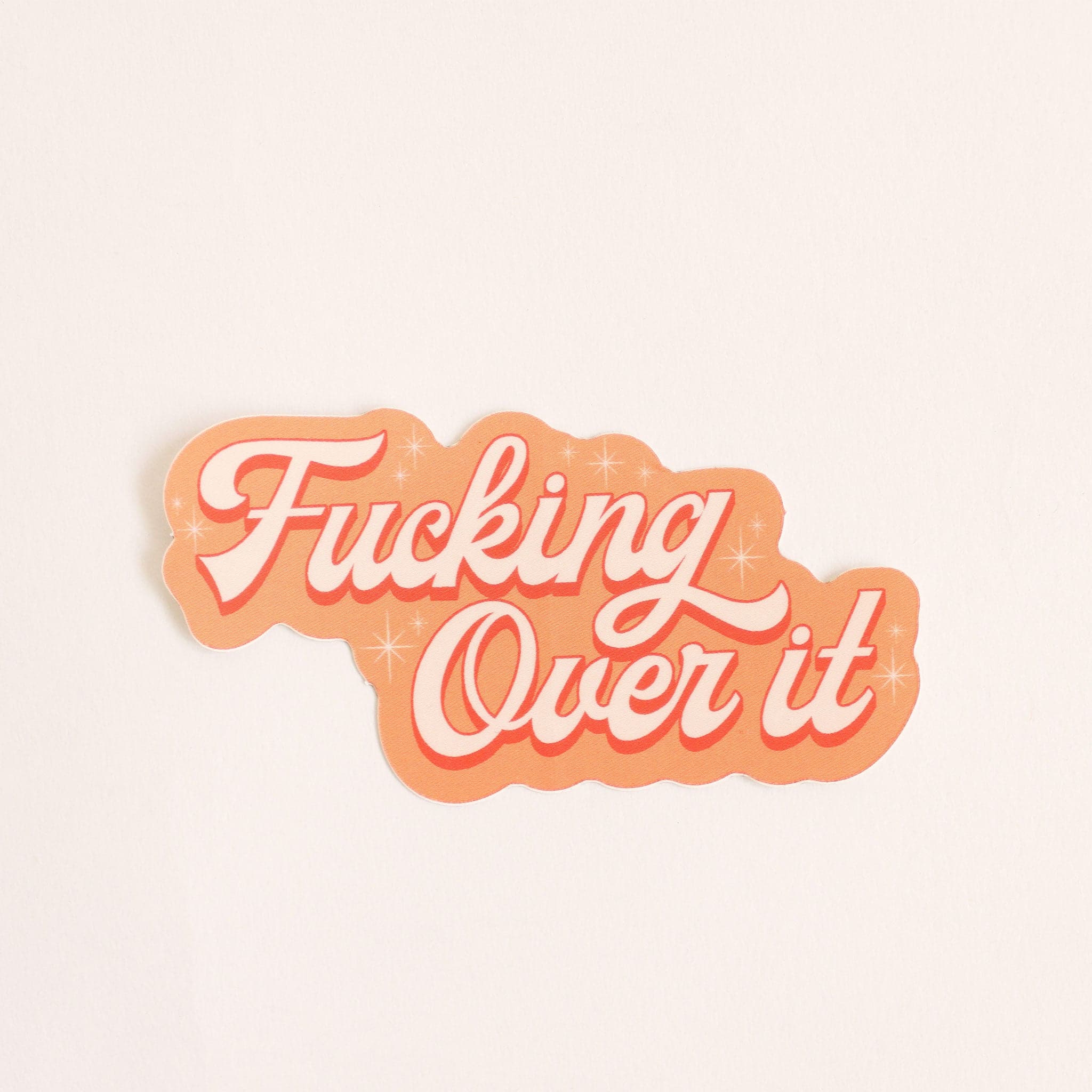 Orange toned sticker that reads &#39;fucking over it&#39; in light cursive lettering. Small twinkle detailing accents around the text. 