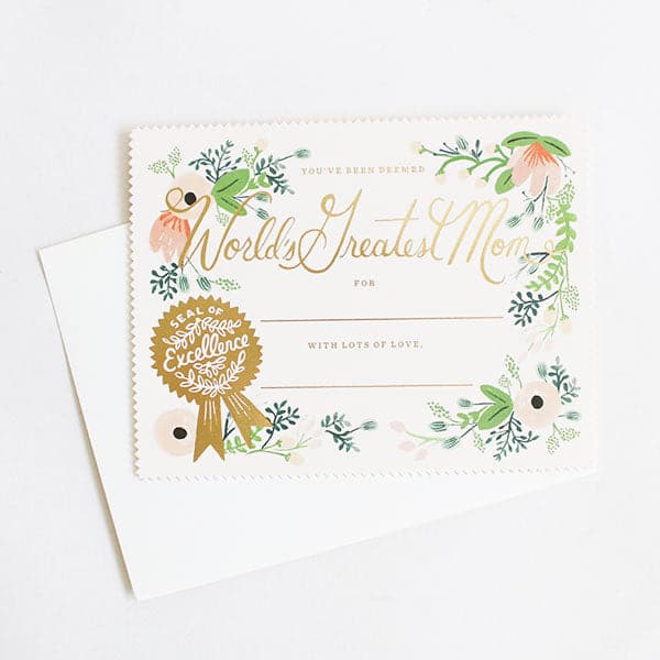 On a white background is a scalloped edge card with florals all around the edge and text that reads, "You've Been Deemed World's Greatest Mom" as well as a place to write "For" and a space underneath "With Lots of Love". It is a flat lay card and also has a gold seal of excellence in the bottom left corner. 