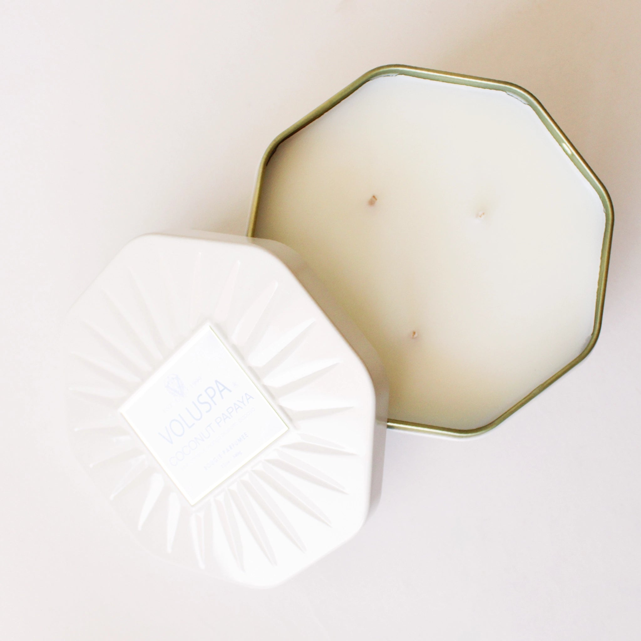On a white background is an octagon tin in a white shade with a white three wick candle inside along with a label on the lid that reads, &quot;Voluspa Coconut Papaya&quot;. 